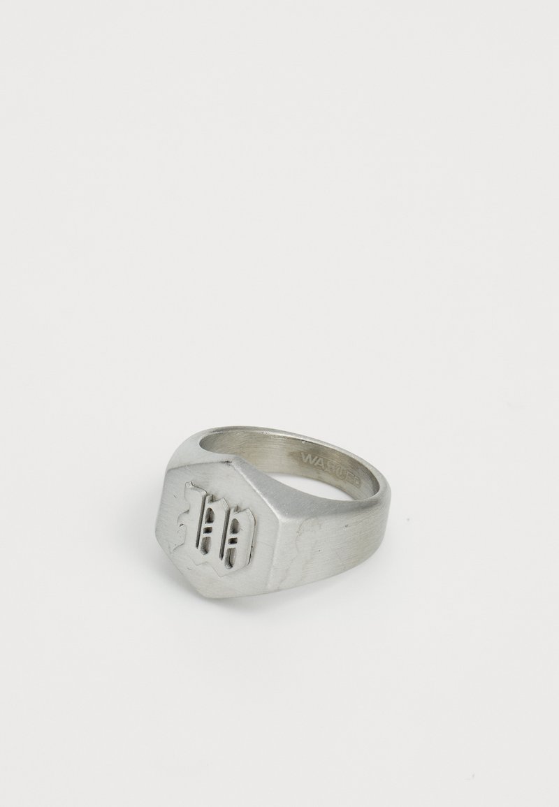 Anel sigilado de metal prateado com forma hexagonal, apresentando um design em relevo que se assemelha a ondas ou riscas na parte da frente. Textura suave.