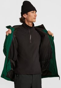 Groene jas met een zwarte voering, open gedragen over een zwarte ritspulli; een outdoor textuur met zichtbare drukknoop- en ritsafsluitingen.