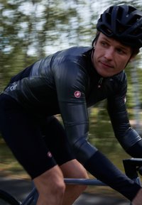 Cyklist i en åtsittande svart långärmad tröja med grå detaljer, i kombination med mörka shorts. Materialet verkar vara slätt och stretchigt.