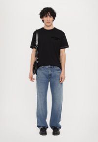 Svart bomullst-skjorte med "JUST CAVALLI" logo, kombinert med lyseblå cowboybukser. Modellen har på seg en svart skulderveske og svarte sko.