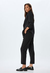 Schwarzes Outfit mit einem lockeren Langarmshirt mit hochgeklappten Ärmelbündchen, passenden schmal zulaufenden Hosen und strukturierten schwarzen Loafers. Schlichtes Design.