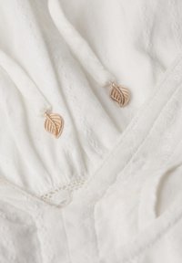 Tejido blanco con delicados detalles de encaje, con dos pequeños charms de hojas en color oro rosa unidos a los cordones. La superficie texturizada añade profundidad.