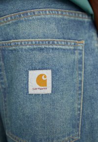 Modra denim tkanina z našitim žepom, na katerem je kvadratna bela nalepka z logotipom Carhartt v oranžni barvi in besedilom v temno rdeči.