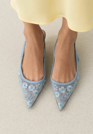 Feet wearing sheer light blue pointed flats with embroidered blue flowers, paired with a yellow hemmed dress.