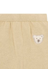 Tessuto lavorato a maglia beige con superficie texturizzata e fascia elastica in vita, con una toppa ricamata a forma di faccia d'orso sul lato sinistro.