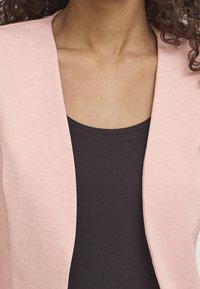 Femme portant un blazer rose clair ouvert sur un haut noir à col rond, montrant uniquement le cou, la poitrine et la partie supérieure du torse.