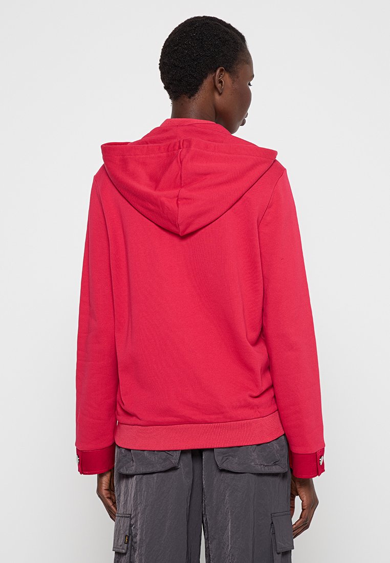 Persona di spalle che indossa una felpa con cappuccio rossa e pantaloni cargo grigi scuri larghi con tasche a pattina.