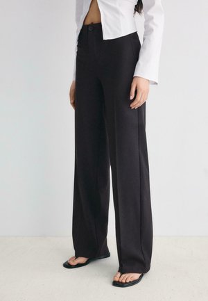 Donna che indossa pantaloni neri a vita alta e gamba larga, giacca corta bianca con bottoni e sandali neri con punta quadrata su sfondo chiaro di pavimento e parete.