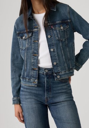 Femme portant une veste en jean délavé moyen sur un haut blanc côtelé et un jean taille haute assorti, main reposant sur la hanche.