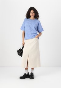Suéter azul claro de malha com mangas curtas, combinado com uma saia bege até aos tornozelos com uma fenda frontal. Sapatos loafers pretos de sola grossa e uma mala com padrão.