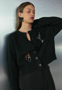 Cardigan negru scurt cu închidere cu nasturi, având un design brodat cu teme de frumusețe pe față. Textură moale cu tiv curb.