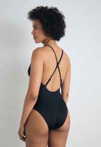 Μαύρο ολόσωμο μαγιό με λεπτές, σταυρωτές  straps και σχεδίαση με χαμηλή πλάτη, κατασκευασμένο από λείο ύφασμα με προσαρμοσμένη εφαρμογή.