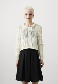 Krämfärgad, stickad hoodie med öppet vävmönster och dragsko, kombinerad med en svart, plisserad, midilång kjol.
