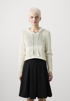Cremefarbener, gestrickter Hoodie mit offener Maschenstruktur und Kordelzügen, kombiniert mit einem schwarzen, gefalteten, knielangen Rock.