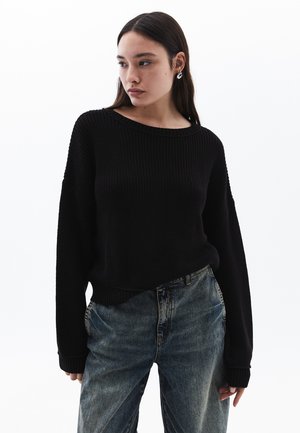 Maglione nero lavorato a maglia con scollatura ampia e vestibilità morbida, abbinato a jeans blu scoloriti. Presenta polsini e orlo a costine per un tocco di texture.