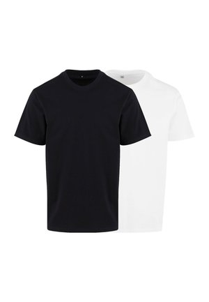 Jeune homme aux cheveux courts portant un t-shirt blanc uni et un pantalon foncé, se tenant devant un fond gris clair.