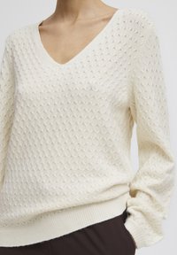 Pull crème avec un col en V et un motif texturé. Dispose de poignets et d'ourlet côtelés, d'un matériau tricoté doux et d'une coupe décontractée.