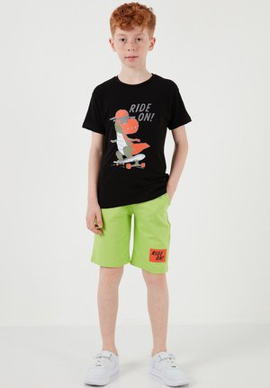 Červenovlasý chlapec v čiernom tričku s potlačou dinosaura na skateboarde a svetlozelených krátkych nohaviciach s nášivkou "RIDE ON!", stojaci s jednou rukou vrecku.