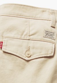 Béžové džínové kalhoty se zadní kapsou, která má bílý knoflík, červenou nášivku Levi's a látkovou etiketu s nápisem "Levi Strauss of California."