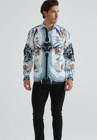 Homme portant une chemise à manches longues avec des motifs floraux et ornementaux complexes en bleu, noir et marron, assortie avec un pantalon noir.