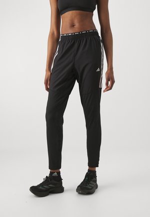 Kvinde iført sorte Adidas løbebukser med hvide striber og en sort sports-bh, stående mod en ensfarvet baggrund.