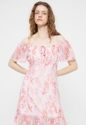 Femme aux cheveux bouclés portant une robe à fleurs rose et rouge, épaules dénudées, posant les mains sur les hanches sur un fond uni.