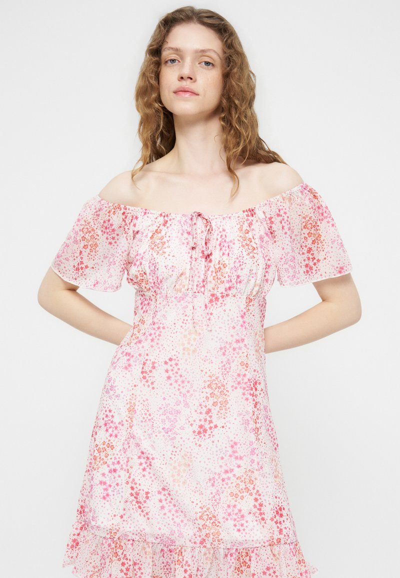 Femme aux cheveux bouclés portant une robe à fleurs rose et rouge, épaules dénudées, posant les mains sur les hanches sur un fond uni.