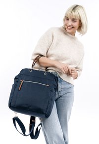 Femme souriante portant un pull beige clair et un jean bleu, tenant un sac à dos bleu marine avec des bretelles en cuir et une poche zippée.