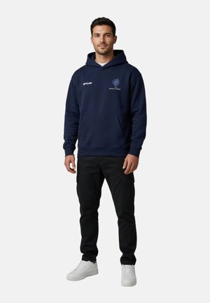 Hombre de pie con una sudadera azul marino con texto blanco y logotipo, pantalones cargo negros y zapatillas blancas sobre un fondo claro y liso.