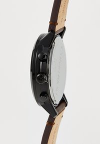Montre noire avec un boîtier rond, bracelet en cuir marron, deux boutons latéraux et une couronne texturée. Le fond est en argent avec un texte gravé.