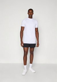 Camiseta deportiva blanca de manga corta con cuello redondo, combinada con pantalones cortos negros. Modelo con zapatillas blancas, mostrando un diseño limpio.