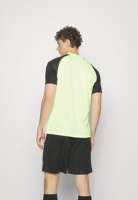 Camiseta atlética de manga corta verde neón con hombros negros y detalles texturizados, combinada con pantalones cortos negros. Presenta un escote en pico.