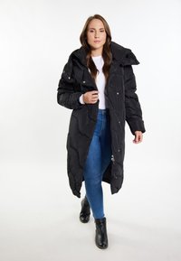 Sort pufferjakke med et langt, quiltet design, stort krave og tryknapper. Bæres over en hvid top og blå jeans, parret med sorte ankelstøvler.