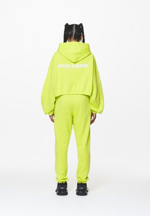 Personne aux cheveux tressés, de dos, portant un survêtement jaune-vert vif avec capuche et des baskets noires sur fond blanc.