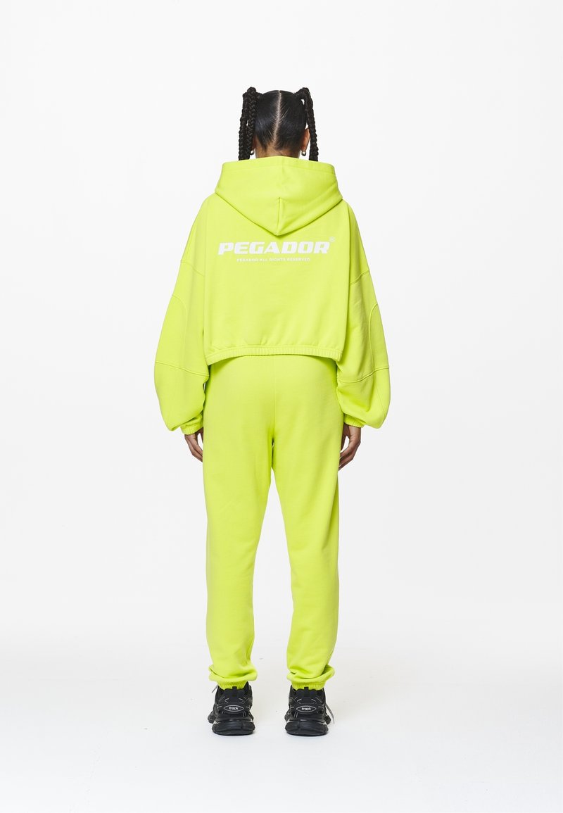 Personne aux cheveux tressés, de dos, portant un survêtement jaune-vert vif avec capuche et des baskets noires sur fond blanc.