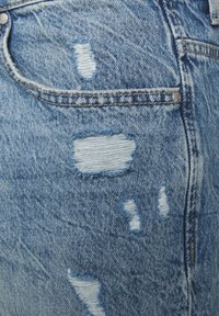 Vaaleansininen kulutettu farkkukangas, jossa repsottavia paikkoja ja erikokoisia repeämiä. Varustettu tavallisella viiden taskun mallilla ja metallisilla koristuksilla.