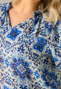 Blusa blu a motivo che presenta un mix di disegni intricati in diverse tonalità di blu su uno sfondo chiaro, con dettaglio di scollatura arricciata.