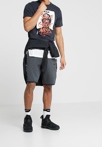 Camiseta negra de Nike con texto gráfico en color naranja, superpuesta sobre una camiseta blanca. Pantalones cortos deportivos grises y zapatillas negras con correas. Calcetines blancos.