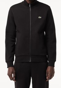 Uomo che indossa una giacca nera con zip e un piccolo logo di coccodrillo verde sul petto, abbinata a pantaloni neri, su sfondo bianco.