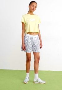 T-shirt amarela corta com um logótipo à frente, combinada com calções cinzentos e brancos às riscas. Footwear com detalhes em prata e meias brancas.