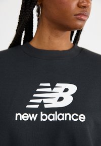 Zwarte sweatshirt met een groot wit "new balance" logo en een gestileerde letter "B." Het materiaal lijkt zacht met een geribbelde halslijn.