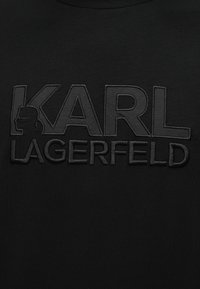 Μαύρο ύφασμα με ανάγλυφα μαύρα κεντημένα γράμματα «KARL LAGERFELD» και τη σιλουέτα ενός ατόμου μέσα στο γράμμα Κ.