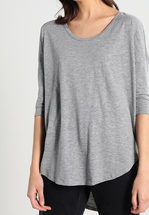 Femme portant un haut gris ample à manches trois-quarts avec un col rond et un pantalon noir sur un fond clair uni.