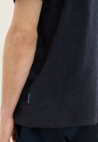Námořnická modrá košile s krátkým rukávem a texturovaným palmovým vzorem, s štítkem na dolní levé straně a uvolněným střihem kolem ramen.