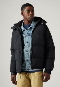 Black puffer jacket with a detachable hood, worn over a light blue denim shirt, featuring button accents and textured materials.