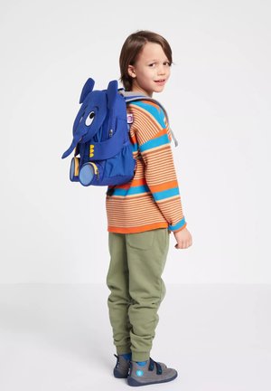 Kind trägt einen gestreiften orange-blauen Pullover mit grüner Hose und grauen Schuhen, trägt einen blauen Rucksack in Elefantenform und schaut zurück.