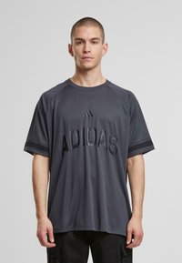 Halli sportlik särk ümmarguse kaelusega ja lühikeste raglanvarrukatega. Omab reljeefset "ADIDAS" teksti ja kontrastset musta triibudetaili.