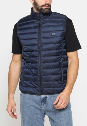 Uomo che indossa un gilet trapuntato blu navy con zip sopra una maglietta nera a maniche corte e jeans azzurri, in piedi con le mani rilassate.