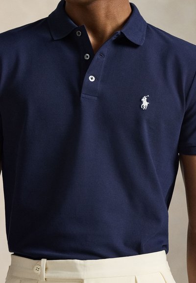 Mies, jolla on päällään tummansininen poolopaita, jonka vasemmassa rinnassa on valkoinen polopelaajan logo, sekä kermaiset housut.