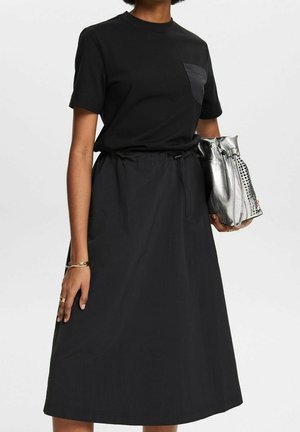 Kobieta w czarnej bluzce z krótkim rękawem i kieszenią na piersi, czarnej spódnicy do kolan, trzymająca srebrną metaliczną, pogniecioną torebkę.